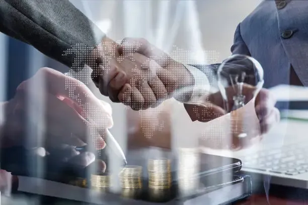 Double exposure of businesspeople shaking hands with coins, documents, and a laptop in the background, symbolizing financial partnership and global business